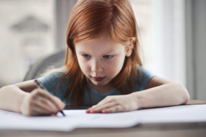 Little redhead girl sitting at a desk and drawing on paper.