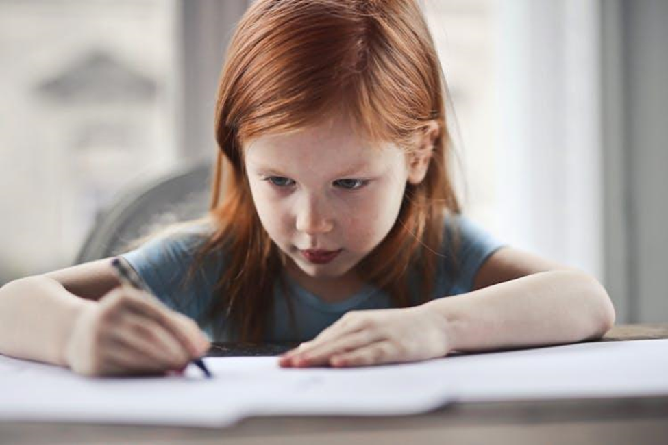 Little redhead girl sitting at a desk and drawing on paper.